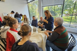 Wokół stołu siedzi osiem osób - trzech mężczyzn i piątka dziewcząt,a jedna kobieta stoi przy stole, na którym leżą elementy do tworzenia rękodzieła.W pomieszczeniu stoją jeszcze cztery kobiety.Jedna ze scian jest całkowicie przeszklona,za nią widać trawnik i drzewa.
