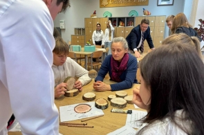 Przy stole siedzi grupa uczniów, którzy trzymają w dłoniach drewniane krążki i oglądają je z bliska. Na blacie leży kilka kolejnych fragmentów drewna, kartki z zadaniami oraz przybory do pisania. Obok nich siedzi dorosła kobieta, patrząca w kierunku krążków. W tle widać inne osoby pracujące w sali oraz szafki