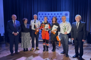 Na zdjęciu znajduje się grupa siedmiu osób, które stoją na scenie. Trzy z nich trzymają w rękach białe róże oraz listy gratulacyjne. Jedna z osób ubrana jest w charakterystyczny pomarańczowy strój ratownika medycznego. Tło sceny jest ciemne, z logotypem medycznym (gwiazda życia) widocznym na plakacie za uczestnikami. 