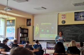 Uczniowie siedzą w sali lekcyjnej, słuchając prelegentki prowadzącej zajęcia w ramach programu „Dermokosmos”. Na ekranie widoczna prezentacja z planetami i logotypami organizatorów. W tle tablica i godło Polski.