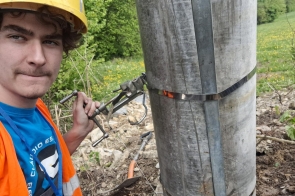 Uczeń w odblaskowej kamizelce i kasku ochronnym pracuje w terenie przy słupie energetycznym, wykorzystując specjalistyczne narzędzie do mocowania obejmy. Zdjęcie dokumentuje praktyczną naukę zawodu w ramach projektu stażowego.