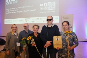 Pięć osób stoi na tle ekranu z logo konferencji REHA FOR THE BLIND IN POLAND. Troje z nich trzyma słoneczniki, jedna osoba trzyma dyplom. Wszyscy są uśmiechnięci, ubrani elegancko. W tle wyświetlone są logotypy partnerów wydarzenia.
