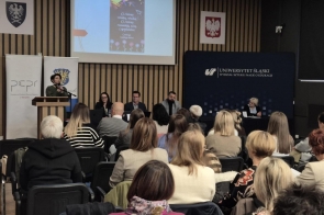 Zdjęcie przedstawia uczestników konferencji zgromadzonych w sali wykładowej Wydziału Sztuki i Nauk o Edukacji Uniwersytetu Śląskiego w Cieszynie. Przy mównicy przemawia Magdalena Bosowska, natomiast za stołem prezydialnym siedzą inni specjaliści. Na ekranie za nimi wyświetlony jest slajd, który zawiera cytat: „Ci, którzy wiedzą, wiedzą. Ci, którzy rozumieją, uczą”. Obok wyświetla się logo Uniwersytetu Śląskiego oraz Powiatowego Centrum Pomocy Rodzinie w Cieszynie. Konferencja dotyczyła tematyki przemocy wobec dzieci, a jej celem była wymiana wiedzy i doświadczeń w walce z tym problemem.