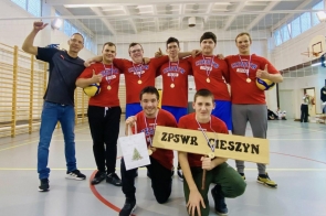 Grupa zawodników w czerwonych koszulkach sportowych pozuje w sali gimnastycznej z medalami na szyjach, uśmiechając się do obiektywu, trzymając piłki siatkarskie oraz tabliczkę z nazwą ZPSWR Cieszyn.