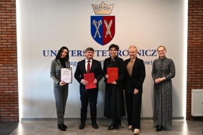 Sześć osób stojących w rzędzie w holu budynku, na tle jasnej ściany z herbem. Trzy osoby trzymają czerwone teczki, jedna osoba trzyma certyfikat w białej oprawie. Uczestnicy ubrani są w stroje formalne. Po bokach ściany widoczne fragmenty ceglanej ściany oraz grzejnik.