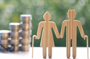 Dwie drewniane sylwetki starszych osób trzymają się za ręce i opierają na laskach, a w tle widać stosy monet oraz rozmytą zieleń.