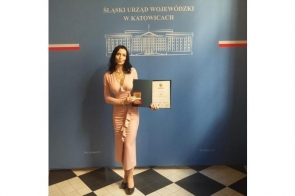 Agata Gacon w eleganckiej, różowej sukni trzyma dyplom i medal na tle ścianki z napisem „Śląski Urząd Wojewódzki w Katowicach”. Uhonorowana w oficjalnym wnętrzu, uśmiecha się z dumą, prezentując swoje wyróżnienie.