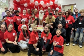 Grupa kilkudziesięciu osób w czerwonych koszulkach z białymi napisami, pozujących wspólnie w sali ozdobionej czerwono-białymi balonami, z osobą ubraną w strój św. Mikołaja stojącą pośrodku, przy stole z tortem.