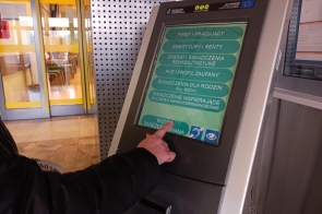 Widoczna ręka osoby w ciemnej kurtce dotyka palcem ekranu dotykowego automatu do obsługi klientów w urzędzie. Na ekranie wyświetlone jest menu wyboru spraw administracyjnych, w tym emerytury, zasiłki, świadczenia rodzinne oraz opcja „wizyta zarezerwowana”. W tle przeszklone drzwi i fragment poczekalni.