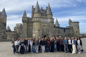 Grupa młodych osób stoi razem przed dużym średniowiecznym zamkiem z kamienia. Budowla ma kilka wysokich, stożkowatych wież i grube mury obronne z wąskimi oknami. Niebo jest jasne, z lekkimi chmurami, a plac, na którym stoją, jest wyłożony brukiem. Ludzie uśmiechają się i pozują do wspólnego zdjęcia.