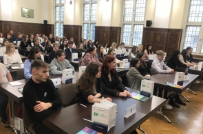 W przestronnej, jasnej sali o wysokich oknach i drewnianych panelach zgromadziła się duża grupa młodych ludzi. Siedzą w rzędach przy stołach, na których ustawiono torby z logo instytucji oraz materiały edukacyjne. Wśród uczestników panuje skupienie, a ich elegancki strój sugeruje oficjalny charakter wydarzenia. Numery na stolikach wskazują na organizację konkursu lub szkolenia.