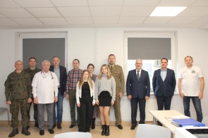 Grupa osób stoi wspólnie w jasnym pomieszczeniu na tle dwóch dużych okien z roletami. Wśród nich są żołnierze w mundurach, lekarz w białym kitlu, mężczyźni w garniturach i kobiety ubrane elegancko. Wszyscy patrzą w obiektyw.