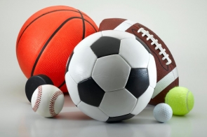 Grafika przedstawia leżące na białym tle, blisko siebie, piłki różnej wielkości. Od lewej na pierwszym planie leży mały czarny krążek hokejowy, mała biała piłka do baseballa z czerwonymi przeszyciami, duża piłka do gry w piłkę nożną w czarno białe łaty, mała biała piłeczka golfowa,mała jasnożółta piłka tenisowa. Na drugim planie duża brązowa piłka do rugby oraz duża pomarańczowa piłka do koszykówki. 