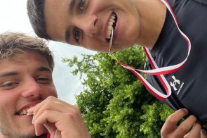Dwóch młodych mężczyzn w sportowych strojach uśmiecha się szeroko, trzymając w ustach medale zawieszone na biało-czerwonych wstążkach. W tle widać drzewa i fragment latarni ulicznej.