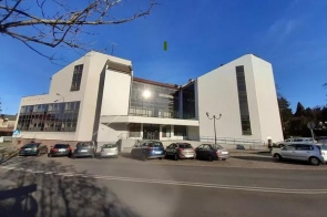Nowoczesny, jasny budynek należący do Banku PKO BP o geometrycznej bryle góruje nad ulicą, odbijając promienie słońca w dużych przeszkleniach. Po jego bokach widoczne są wysokie, wąskie okna. Przed obiektem rozciąga się parking, gdzie zaparkowanych jest kilka samochodów. Po prawej stronie widać podjazd dla osób z niepełnosprawnościami. Nad wszystkim rozciąga się bezchmurne, błękitne niebo, a w oddali dostrzegalna jest zieleń drzew.