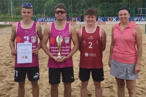 Cztery osoby stoją na boisku do siatkówki plażowej przed siatką z logo ORLEN. Dwóch chłopaków trzyma dyplom i puchar, a w tle widoczne jest piaszczyste boisko i drzewa.