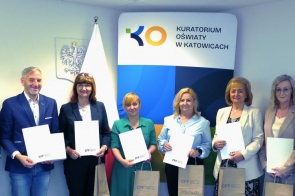Siedem osób w eleganckich strojach stoi w rzędzie, trzymając białe teczki z logo „CFF”. W tle widoczna flaga Polski, godło oraz ścianka z napisem „Kuratorium Oświaty w Katowicach”.