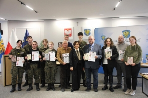 Grupa osób stoi wspólnie na sali sesyjnej, prezentując trzymane w rękach dyplomy i statuetki. W centralnej części pierwszego rzędu znajduje się Starosta Cieszyński Janina Żagan, elegancko ubrana w czarne spodnie i czarną marynarkę. Towarzyszą jej młodzi uczestnicy w mundurach oraz dorośli laureaci wyróżnień. W tle widoczne są flagi oraz godła.