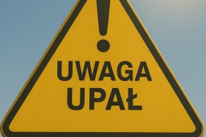 Żółty, trójkątny znak ostrzegawczy z czarnym obramowaniem i napisem „Uwaga upał” pod dużym wykrzyknikiem, ustawiony na tle błękitnego nieba z widocznym w rogu słońcem.