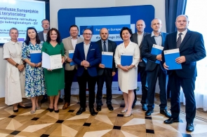 Grupa kobiet i mężczyzn w eleganckich strojach stoi na tle tablic i ekranów z logotypami Funduszy Europejskich. Część osób trzyma teczki lub dokumenty, uśmiechając się do obiektywu, co podkreśla oficjalny i uroczysty charakter spotkania.