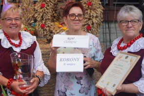 Na zdjęciu widzimy trzy kobiety, które reprezentują Koło Gospodyń Wiejskich Czaniec. Stoją one razem, dumnie prezentując swoje nagrody. Dwie z kobiet mają na sobie tradycyjne stroje ludowe, składające się z białych bluzek z koronkowymi kołnierzykami oraz ciemnoczerwonych gorsetów. Kobiety te noszą także korale w intensywnym czerwonym kolorze. Kobieta po lewej stronie trzyma w rękach szklany puchar z napisem, który wskazuje na zajęcie miejsca w konkursie. Kobieta w środku trzyma tabliczkę z napisem "KGW CZANIEC" oraz drugą tabliczkę z napisem "KORONY II miejsce", wskazującą na zajęcie drugiego miejsca w konkursie w kategorii "korony". Kobieta po prawej stronie trzyma dyplom oprawiony w ramkę, który również potwierdza zdobycie nagrody. W tle widoczne są dożynkowe wieńce wykonane z kłosów zbóż i ozdobione kolorowymi kwiatami.