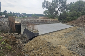 Zdjęcie przedstawia plac budowy, na którym trwa budowa nowego mostu w Kończycach Małych. W centralnej części fotografii widoczna jest konstrukcja betonowa, która stanowi podstawę nowo powstającego mostu. Betonowa płyta mostowa jest już wylana i widać zbrojenie wystające ponad jej powierzchnię, co wskazuje, że prace budowlane są w toku. Po obu stronach mostu widoczne są tymczasowe drewniane barierki zabezpieczające. W tle widoczne są drzewa oraz budynki mieszkalne, co pokazuje, że budowa odbywa się w pobliżu obszarów zamieszkałych. Na zdjęciu panuje pochmurna pogoda, co tworzy stonowaną kolorystykę obrazu.