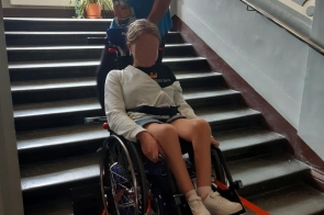 Na zdjęciu widać mężczyznę, który pomaga osobie na wózku inwalidzkim pokonać schody za pomocą schodołazu. Mężczyzna stoi za wózkiem, trzymając uchwyty urządzenia, podczas gdy osoba na wózku siedzi z zapiętymi pasami bezpieczeństwa. Schodołaz jest umieszczony na schodach i umożliwia bezpieczne transportowanie osoby na wózku w górę lub w dół po schodach. W tle widoczna jest ściana z poręczą, a przez okno wpada światło, oświetlając wnętrze budynku. Na parapecie znajduje się roślina doniczkowa.