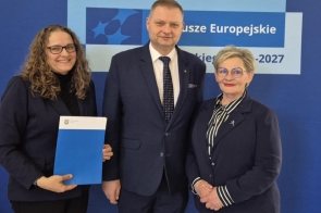 Trzy osoby w eleganckich strojach (od lewej Agnieszka Baszczyńska, dyrektor PCPR w Cieszynie, od prawej Beata Macura, Członkini Zarządu Powiatu Cieszyńskiego, w środku Rafał Adamczyk, Członek Zarządu Województwa Śląskiego) stoją na tle niebieskiej ścianki z napisem „Fundusze Europejskie 2021–2027”. Agnieszka Baszczyńska trzyma teczkę z logo województwa śląskiego. Wszyscy uśmiechnięci, pozują do wspólnego zdjęcia.