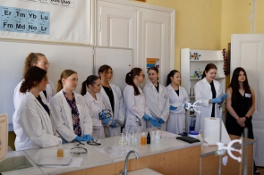 Grupa młodych kobiet w białych fartuchach laboratoryjnych stoi w sali chemicznej, przygotowana do pokazu lub prezentacji. Na ścianie widoczna tablica z układem okresowym pierwiastków i modele cząsteczek.