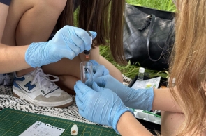 Uczennice w rękawiczkach ochronnych wykonują doświadczenie chemiczne w plenerze, dodając ciecz z pipety do probówki. W tle widać trawę, torbę i zestaw do badań terenowych. Na dole leży instrukcja i mata pomiarowa.