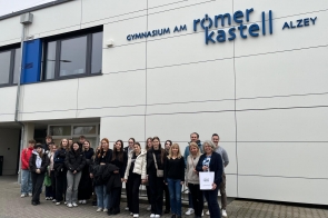 Grupa osób stoi przed budynkiem szkoły o nazwie "Gymnasium am Römerkastell Alzey". W tle widoczna jest biała elewacja z dużymi oknami. Osoby są uśmiechnięte i różnie ubrane, niektóre trzymają torby.