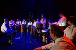 Grupa osób siedzi w półkolu na scenie, grając na bębnach, większość ubrana jest w białe koszule z muszkami, a jedna osoba w różowej bluzce stoi pośrodku i również gra, scena oświetlona jest niebieskim i czerwonym światłem