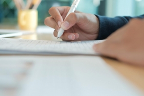 Dłoń trzymająca biały długopis pisze na kartce papieru, widoczny fragment blatu oraz rozmyte tło z przedmiotami biurowymi.