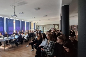 Uczennice i uczniowie uważnie słuchają podczas spotkania w jasnej sali z niebieskimi zasłonami, siedząc przy stołach i na krzesłach ustawionych w kilku rzędach; w tle widać bibliotekę i szkolny projektor.