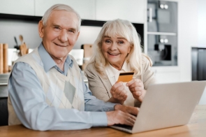 Starszy mężczyzna i starsza kobieta siedzą przy biurku z laptopem; kobieta trzyma kartę płatniczą, mężczyzna trzyma ręce na klawiaturze.