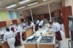 Grupa uczniów w białych strojach kucharskich i bordowych fartuchach pracuje w szkolnej pracowni gastronomicznej. Część osób wyrabia ciasto przy blatach, inni przygotowują składniki i korzystają z piekarników oraz kuchenek gazowych. Pomieszczenie wyposażone jest w drewniane szafki, metalowe blaty i oświetlenie sufitowe.