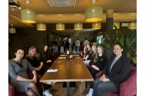 Grupa uczniów w eleganckich strojach hotelarskich ćwiczy przy recepcji. W tle widoczna jest tablica z nazwą obiektu, a atmosfera spotkania jest pełna zaangażowania i uśmiechów.