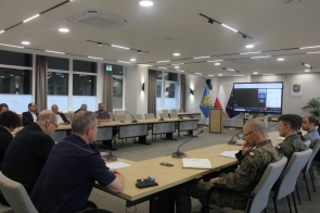 Uczestnicy spotkania – przedstawiciele samorządu, służb mundurowych i instytucji – siedzą przy stołach w sali konferencyjnej. Na ekranie trwa wideokonferencja. W tle widoczne flagi: Polski, UE i Powiatu Cieszyńskiego.