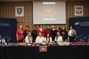 Grupa kilkunastu osób pozuje razem do zdjęcia. Z tyłu, na ekranie projektora, widoczny jest plakat 16. Cieszyńskiej Wampiriady. W pierwszym rzędzie za stołem siedzą młode osoby o, a za nimi stoi grupa kobiet w tym w odświętnych, czerwonych i zimowych stylizacjach. Po bokach widoczne są elementy scenografii z logo Uniwersytetu oraz godła.