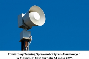 Syrena alarmowa zamontowana na słupie, skierowana w lewą stronę, na tle czystego, niebieskiego nieba. W dolnej części obrazka znajduje się informacja o powiatowym treningu syren alarmowych w Cieszynie.