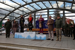 Trzy kobiety stoją na podium zwyciężczyń zawodów sportowych w Ustroniu; otrzymują róże i upominki. Obok nich stoją uśmiechnięci przedstawiciele wojska i władz lokalnych w otwartej, zadaszonej przestrzeni miejskiej.