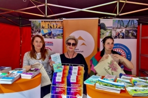 Trzy kobiety – od lewej: Aneta Brożkowska, główna specjalistka w Biurze ds. Kultury, Sportu, Turystyki i Promocji Powiatu, Beata Macura, Członkini Zarządu, Barbara Jurkiewicz, Kierowniczka Biura ds. Kultury, Sportu, Turystyki i Promocji Powiatu, stoją za stoiskiem pełnym kolorowych ulotek i broszur. Uśmiechają się, trzymając torby i materiały promocyjne. Tło ozdobione jest plakatami z motywami turystycznymi. Jasne, soczyste kolory tworzą przyjazną atmosferę.
