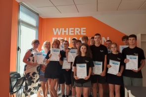 Grupa młodych osób wraz z nauczycielkami pozuje do zdjęcia w jasnym pomieszczeniu z pomarańczową ścianą z napisem. Wszyscy trzymają w rękach certyfikaty, uśmiechając się do kamery.