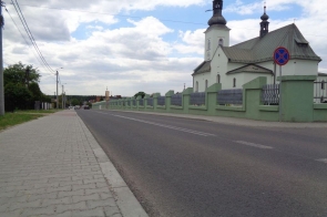 Droga asfaltowa biegnie obok kościoła z dwiema wieżami, otoczonego zielonym murem. W tle widać domy i drzewa pod pochmurnym niebem. Po lewej stronie drogi znajduje się chodnik z betonowych płyt.
