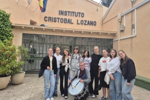 Grupa młodych osób stoi przed budynkiem z napisem "Instituto Cristobal Lozano". W centrum grupy znajduje się osoba z bębnem. W tle widać rośliny w donicach i flagi na dachu. Uczestnicy są uśmiechnięci i ubrani na sportowo.