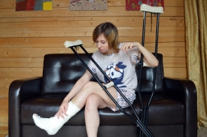 Kobieta w piżamie siedzi na ciemnobrązowej skórzanej kanapie, opierając się o nią i trzymając kule ortopedyczne. Ma nogę w gipsie, którą delikatnie dotyka. Tło stanowi drewniana ściana z trzema kolorowymi obrazami, a po prawej stronie widać beżową zasłonę.