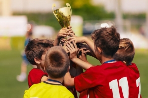 Grupa małych chłopców na boisku unosi razem złoty puchar. Czerwone koszulki piłkarskie, numery na plecach, medale na szyjach; jeden w żółtej bluzie. W tle rozmyta murawa i jasny dzień, euforia po wygranej. Dłonie na pucharze, miny skupione. W górze!