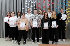 Grupa młodych osób stoi na tle szarej kotary ozdobionej kolorowymi motylami. Trzymają teczki z logo ZSTiO. Ubrani elegancko i półformalnie, ustawieni w dwóch rzędach, patrzą poważnie lub z lekkimi uśmiechami.