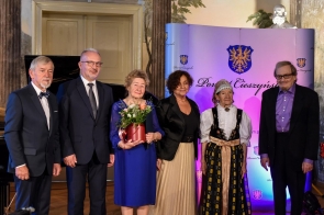 Sześć osób stojących w szeregu. Od lewej: od lewej Stanisław Kubicius, Mieczysław Szczurek, prof. Heska-Kwaśniewicz, Janina Żagan, Helena Wróbel, Adam Makowicz. Dwie kobiety znajdują się po środku, jedna z nich trzyma w rękach czerwony wazon z kwiatami. Kobieta po prawej stronie ubrana jest w tradycyjny strój ludowy. Pozostałe osoby mają na sobie eleganckie stroje, głównie garnitury. W tle znajduje się niebieska ścianka z herbem oraz napisem "Powiat Cieszyński".
