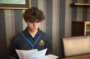 Chłopak z identyfikatorem na niebieskiej smyczy siedzi przy stole i przegląda dokumenty. Przed nim stoi laptop, a w tle widoczna jest kartka z informacją o stażu uczniowskim oraz obraz na ścianie.